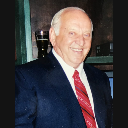 A close-up photo of John Mikus Jr. smiling while dressed in a black suit and red tie