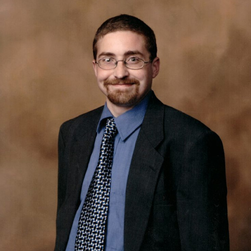 A professional portrait photo of Joshua "Josh" "Knuch" Knuchel smiling and dressed in a black suit and tie