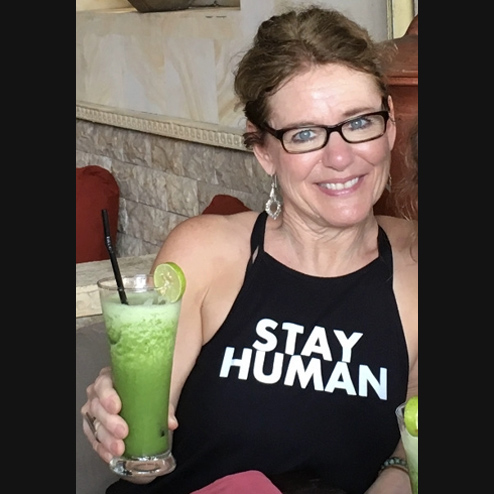 A close-up photo of Kim M. ONeill smiling while wearing a "Stay Human" tshirt and drinking a green juice