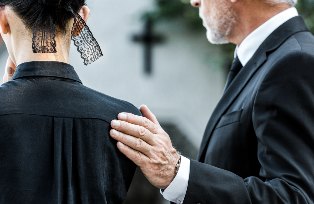 A gentleman dressed in black with his hand on the back of a woman dressed in black at a funeral setting