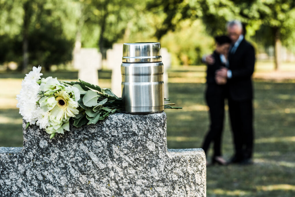 A couple embracing in the distance with an urn resting on a cemetery rock and flowers in focus at the front