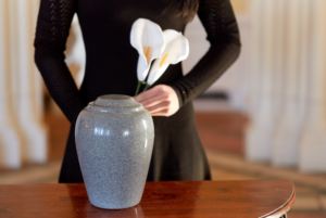 A woman standing in the background dressed in black holding a lily in front of an urn to represent Funeral Trends of 2024 from Preplanning to Cremation Rates