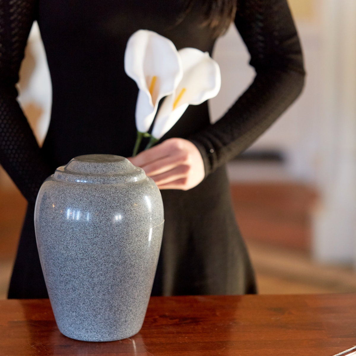 A woman standing in the background dressed in black holding a lily in front of an urn to represent Funeral Trends of 2024 from Preplanning to Cremation Rates