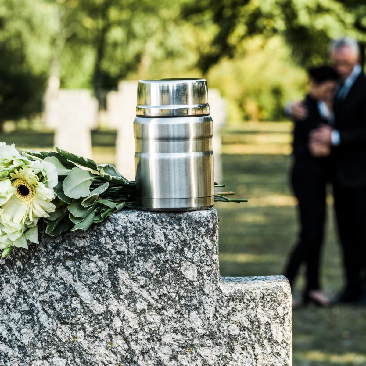 A couple embracing in the distance with an urn resting on a cemetery rock and flowers in focus at the front